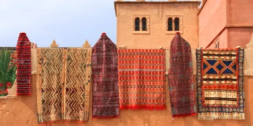 Row of traditional Berber rugs hanging on an ochre wall in Morocco, each showcasing intricate Berber symbols and vibrant geometric patterns.